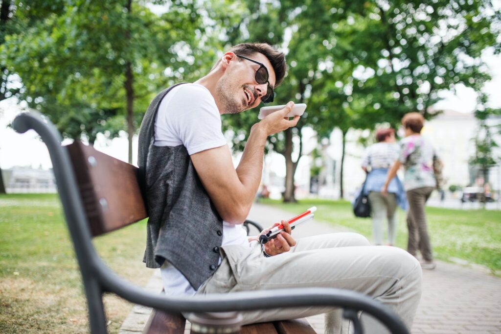 Young blind man with smartphone sitting on bench in park in city
